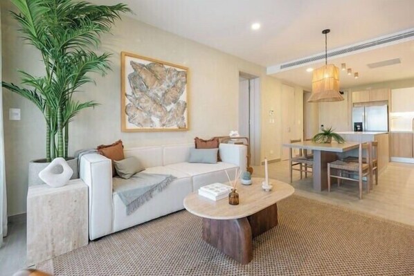 A modern living room with a white sofa, a coffee table, a large plant, and a painting on the wall.