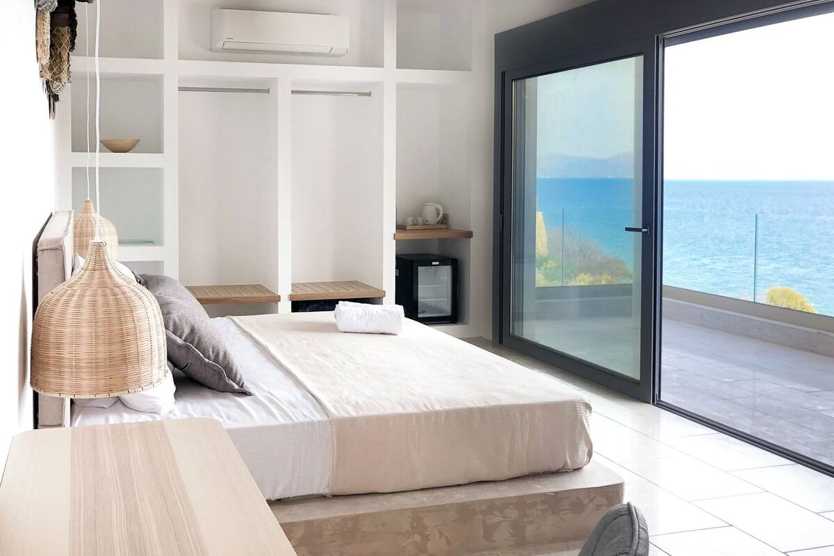 A bedroom with a bed, bedside tables, a small heater, a window with a view of the sea, and a balcony.