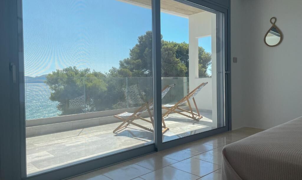 Een kamer met een grote schuifglazen deur die uitkomt op een balkon met uitzicht op de zee en bomen.