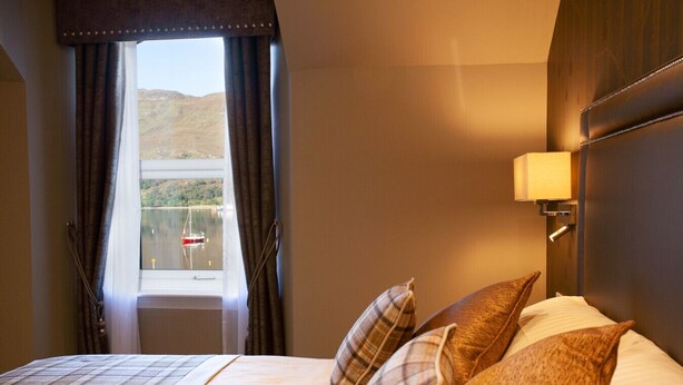 Ein Hotelzimmer mit Bett, einem Fenster mit Vorhängen, Blick auf einen See und Berge sowie einer Wandlampe.