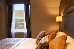 Une chambre d’hôtel avec un lit, une fenêtre avec des rideaux, une vue sur un lac et des montagnes, et une lampe fixée au mur.