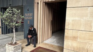 A man sitting outside a building labeled 'ALBERT LUXURY II' with a potted tree and a tiled floor.