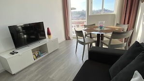 Un salon moderne avec un téléviseur à écran plat posé sur une console blanche, un canapé noir, une table à manger avec des chaises, et une vue sur la ville grâce à de grandes fenêtres.