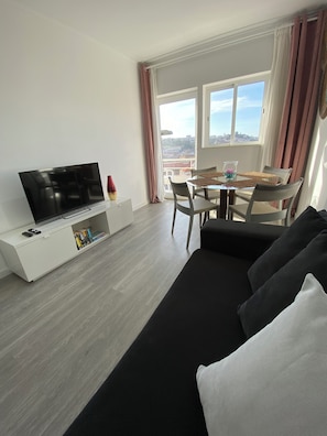 A modern living room with a flat-screen TV on a white console, a black sofa, a dining table with chairs, and a view of the city through large windows. - Spectacular view of Porto and Douro-Free Parking (Vila Nova de Gaia)