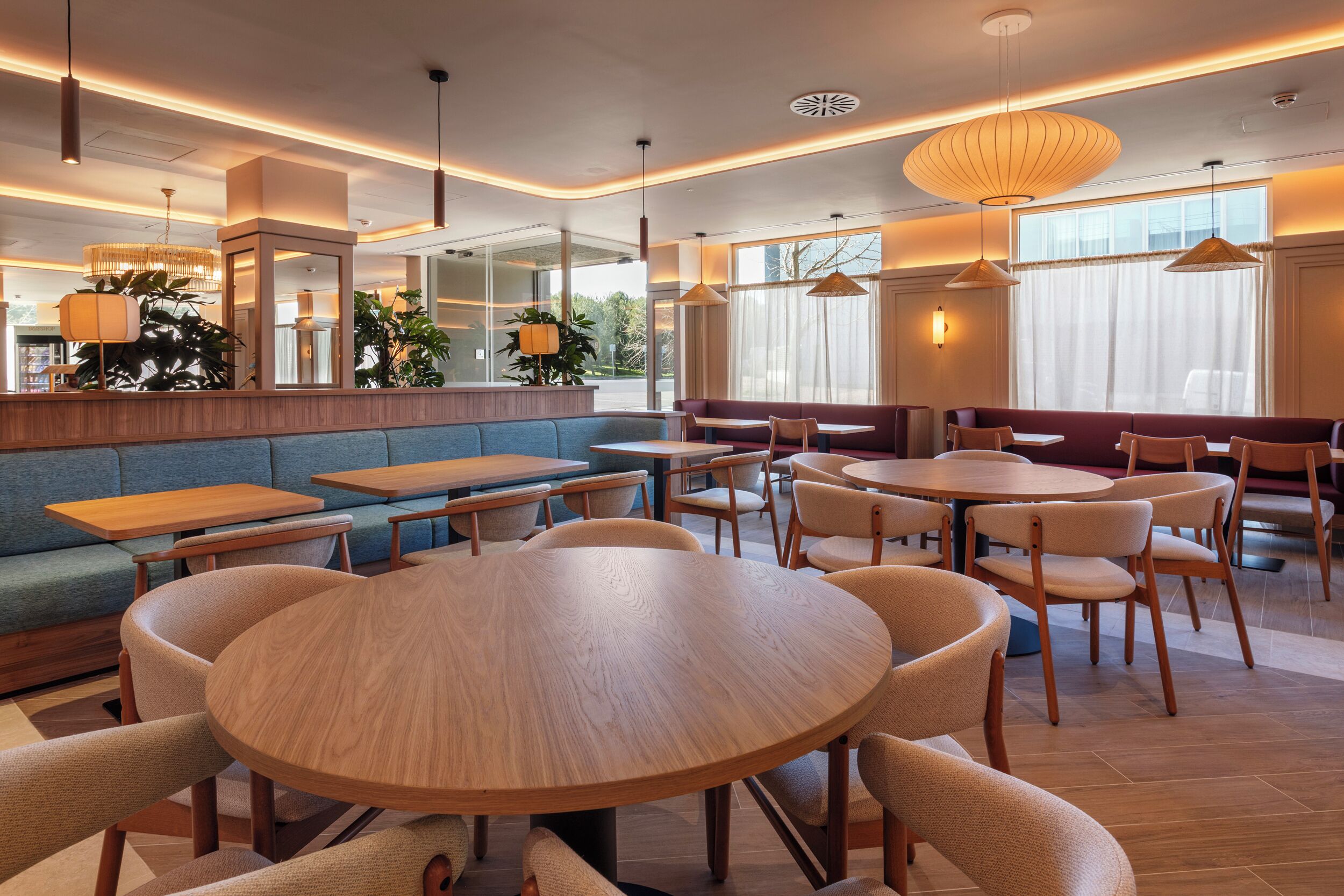 Ein modernes Restaurant mit runden Holztischen, gepolsterten Sitzplätzen, Pendelleuchten und großen Fenstern.