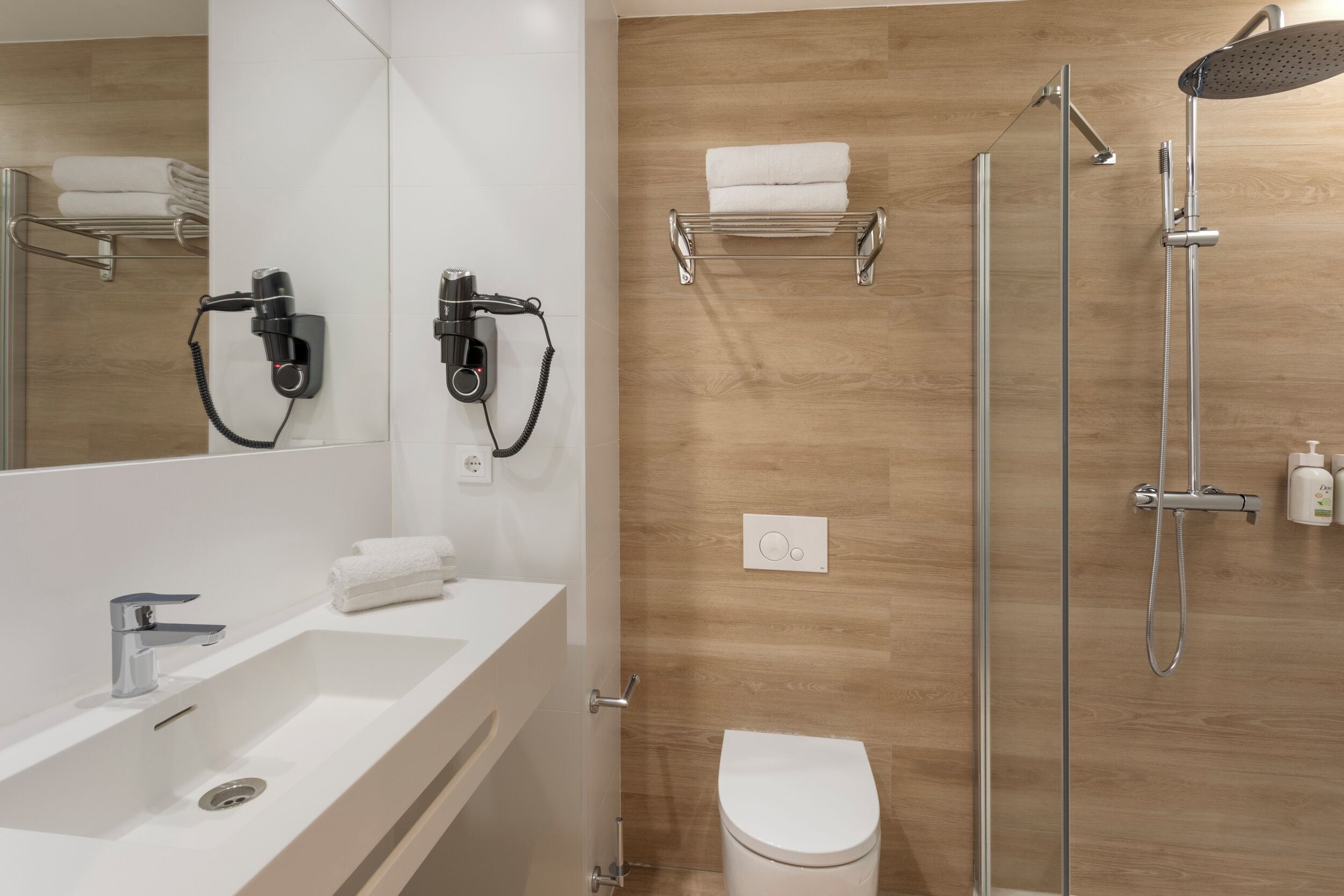 Un baño moderno con un lavabo blanco, dos secadores de pelo montados en la pared, una ducha con mampara de vidrio y un inodoro.