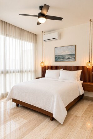 Une chambre d’hôtel avec un lit, un ventilateur de plafond, un appareil de climatisation, un tableau représentant la mer et deux lampes fixées au mur.