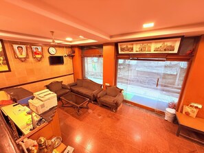 Eine gemütliche Hotellobby mit einer Sofagarnitur, einem Couchtisch und einem Schreibtisch mit verschiedenen Gegenständen. An der Wand hängen gerahmte Bilder und ein großes Fenster mit Blick auf die Straße.