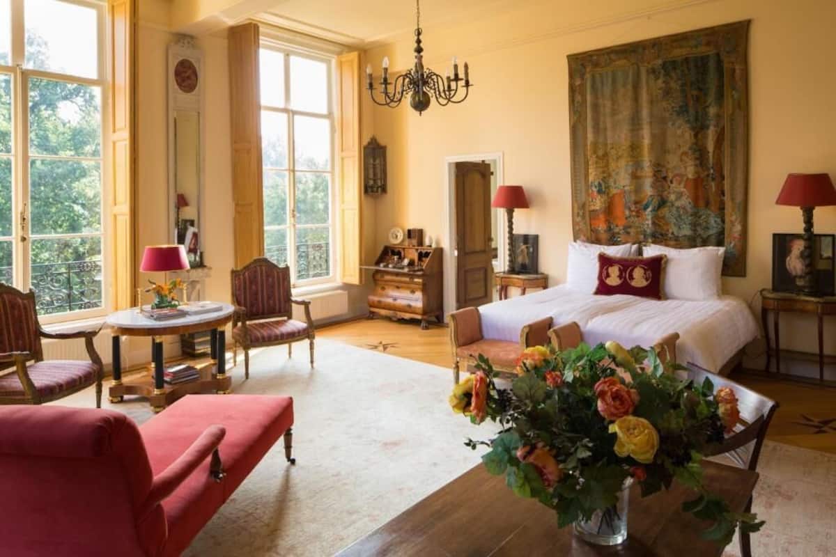 A spacious room with a large bed, a sitting area with a red armchair, a table with a vase of flowers, and a large window with a view of trees.