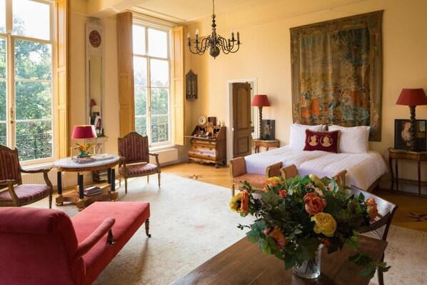 A spacious room with a large bed, a sitting area with a red armchair, a table with a vase of flowers, and a large window with a view of trees. - Kasteel Sterkenburg (Driebergen)