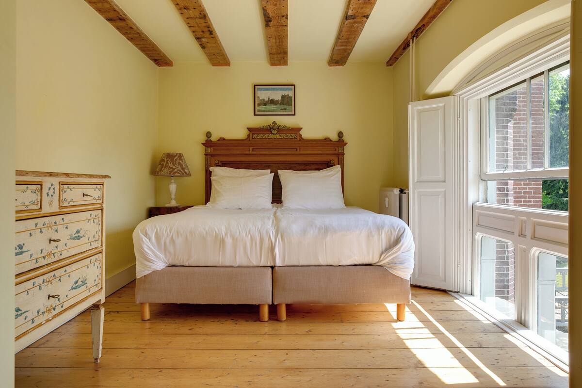 A bedroom with a wooden bed, a dresser with drawers, a lamp, and a window with a view of greenery.
