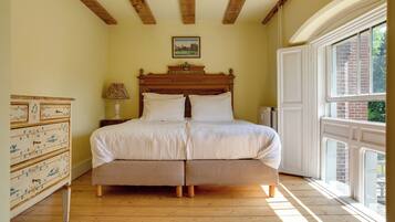 A bedroom with a wooden bed, a dresser with drawers, a lamp, and a window with a view of greenery.