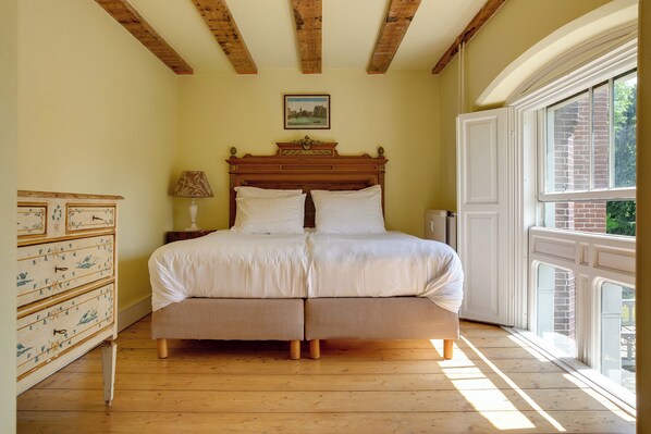 A bedroom with a wooden bed, a dresser with drawers, a lamp, and a window with a view of greenery. - Kasteel Sterkenburg (Driebergen)