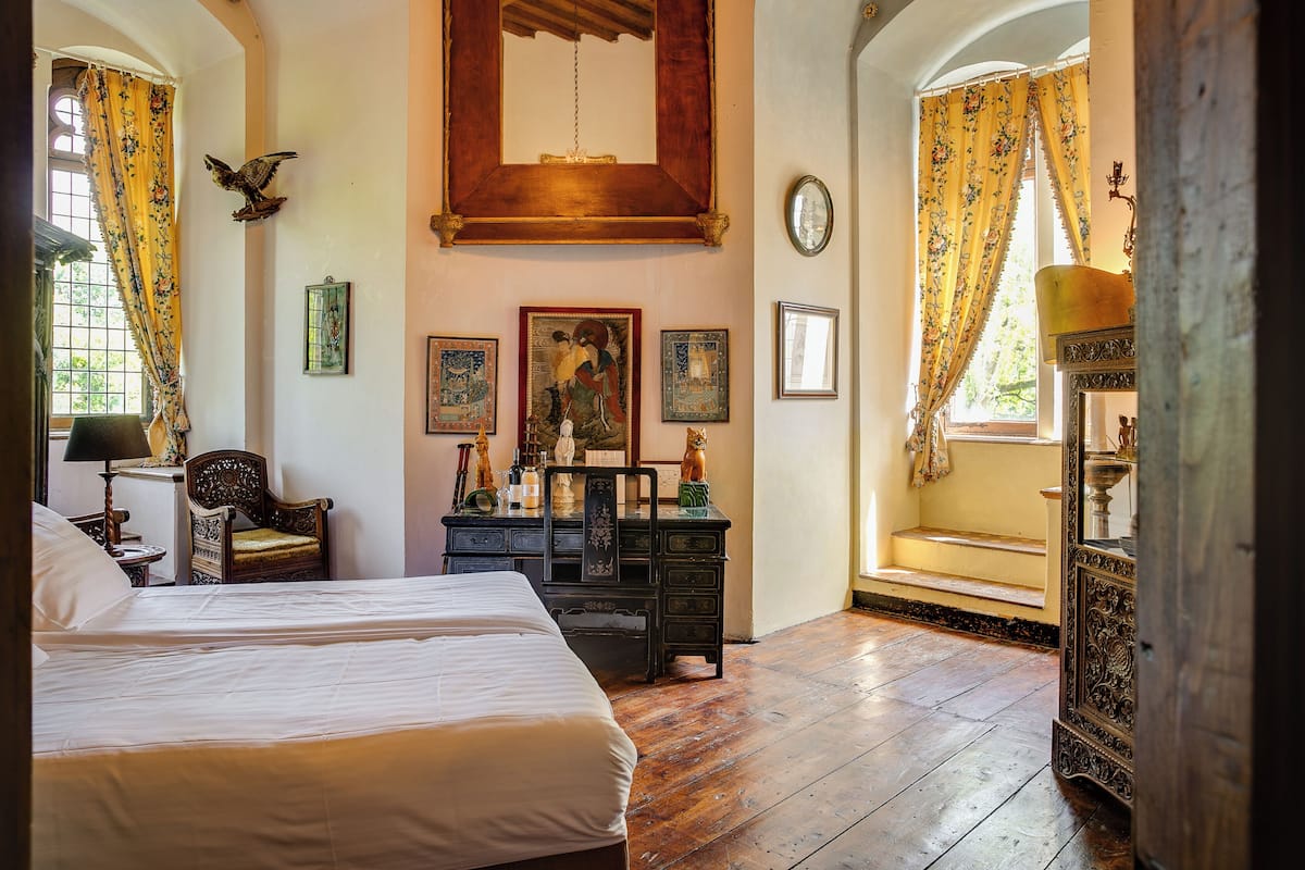 A room with two beds, wooden floors, a large mirror, and a window with yellow curtains.
