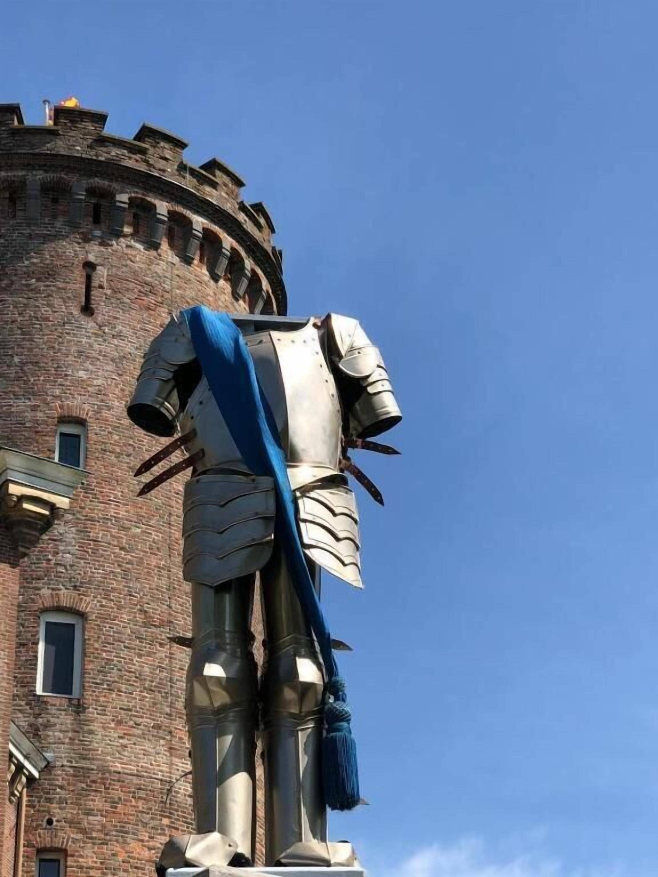 A statue of a knight in armor with a blue sash, standing in front of a brick tower.