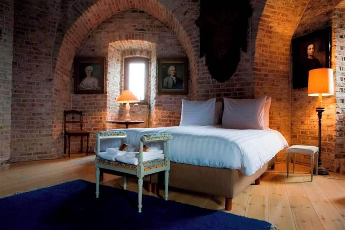 A bedroom with a brick arched ceiling, a bed with white linens, two bedside tables with lamps, and a chair with a folded towel.