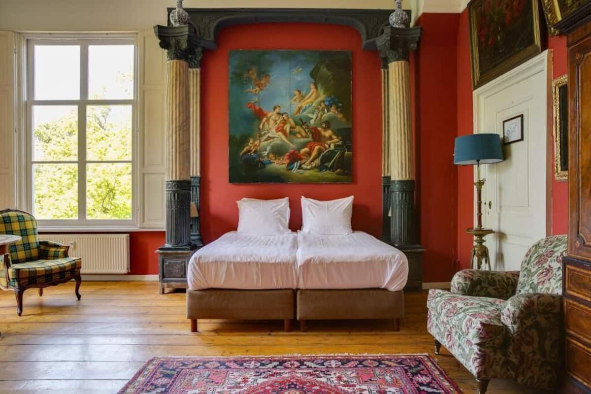 A room with a large bed, two chairs, a painting on the wall, and a window.