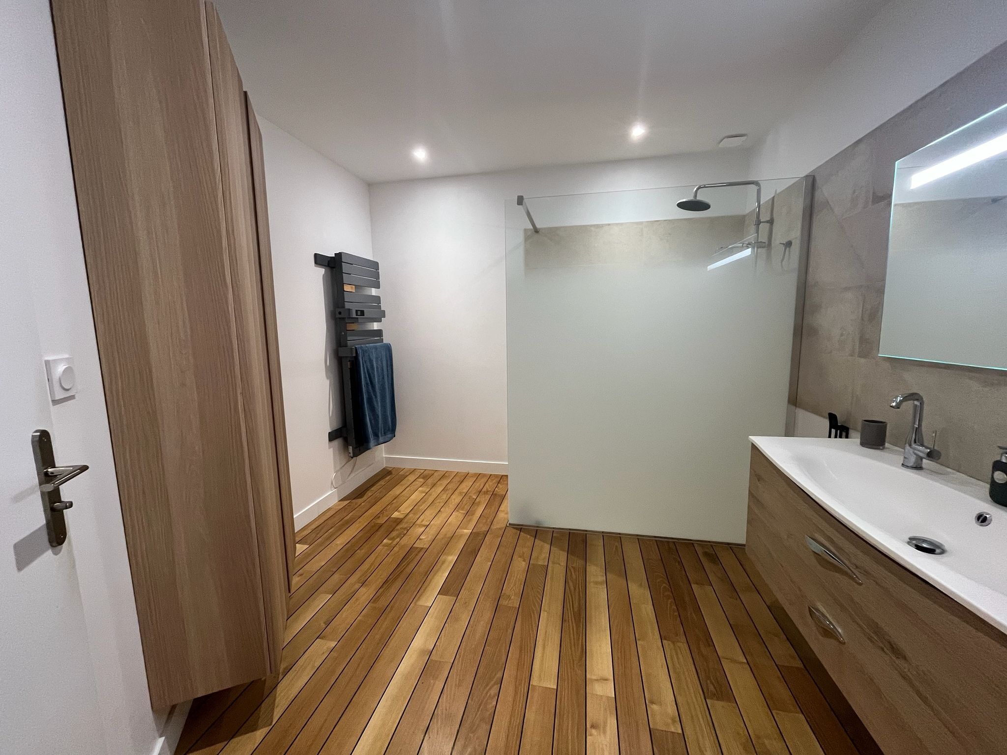 A modern bathroom with a wooden floor, a white sink, a mirror, and a shower.
