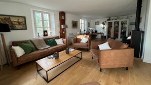 Een ruime woonkamer met een bruine leren bank, een houten salontafel en een bruine fauteuil. Er is een groot raam dat natuurlijk licht binnenlaat, een houten vloer en een open haard.
