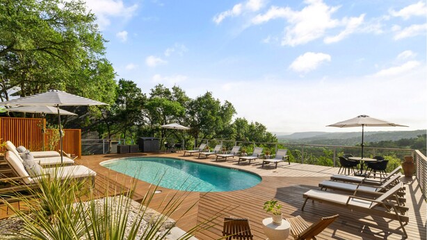 Une piscine entourée d’une terrasse en bois et de verdure, avec des chaises longues et des parasols.