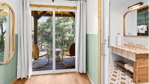 Une pièce dotée d’un plancher en bois, d’une grande porte-fenêtre coulissante donnant sur un espace extérieur aménagé avec des chaises, d’un plafond en bois et d’une salle de bain avec un lavabo et un miroir.