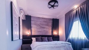 A modern bedroom with a large bed, a headboard with built-in lighting, a wall-mounted air conditioner, and a large window with curtains. - Valyron Homes (Accra)