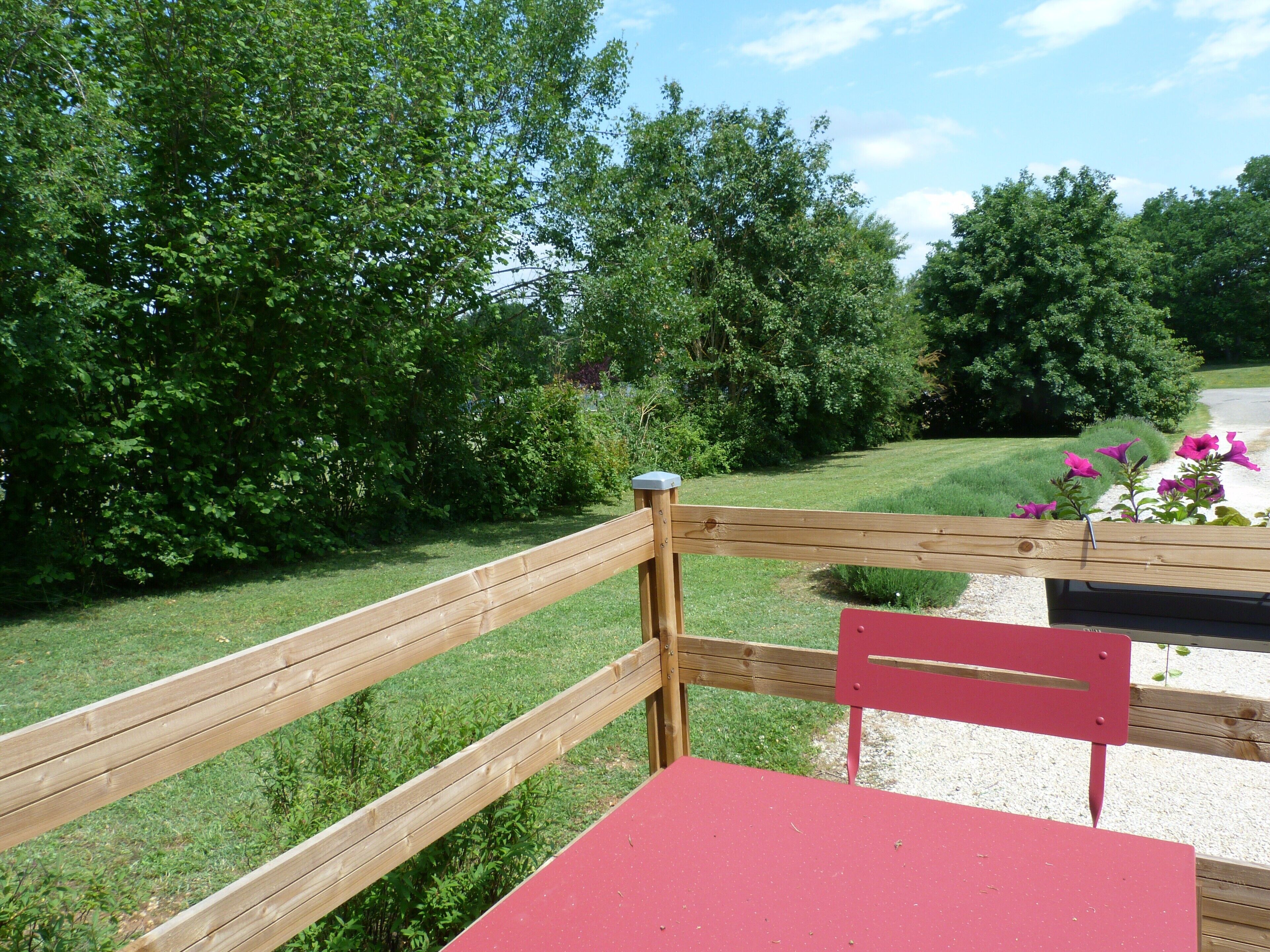 A wooden deck with a red table and chairs, surrounded by lush greenery and trees.
