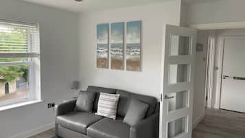 Un salon moderne avec un canapé en cuir gris, un coussin décoratif à motifs et une vue sur l’extérieur par une fenêtre. Trois tableaux sont accrochés au mur et représentent une scène de plage.