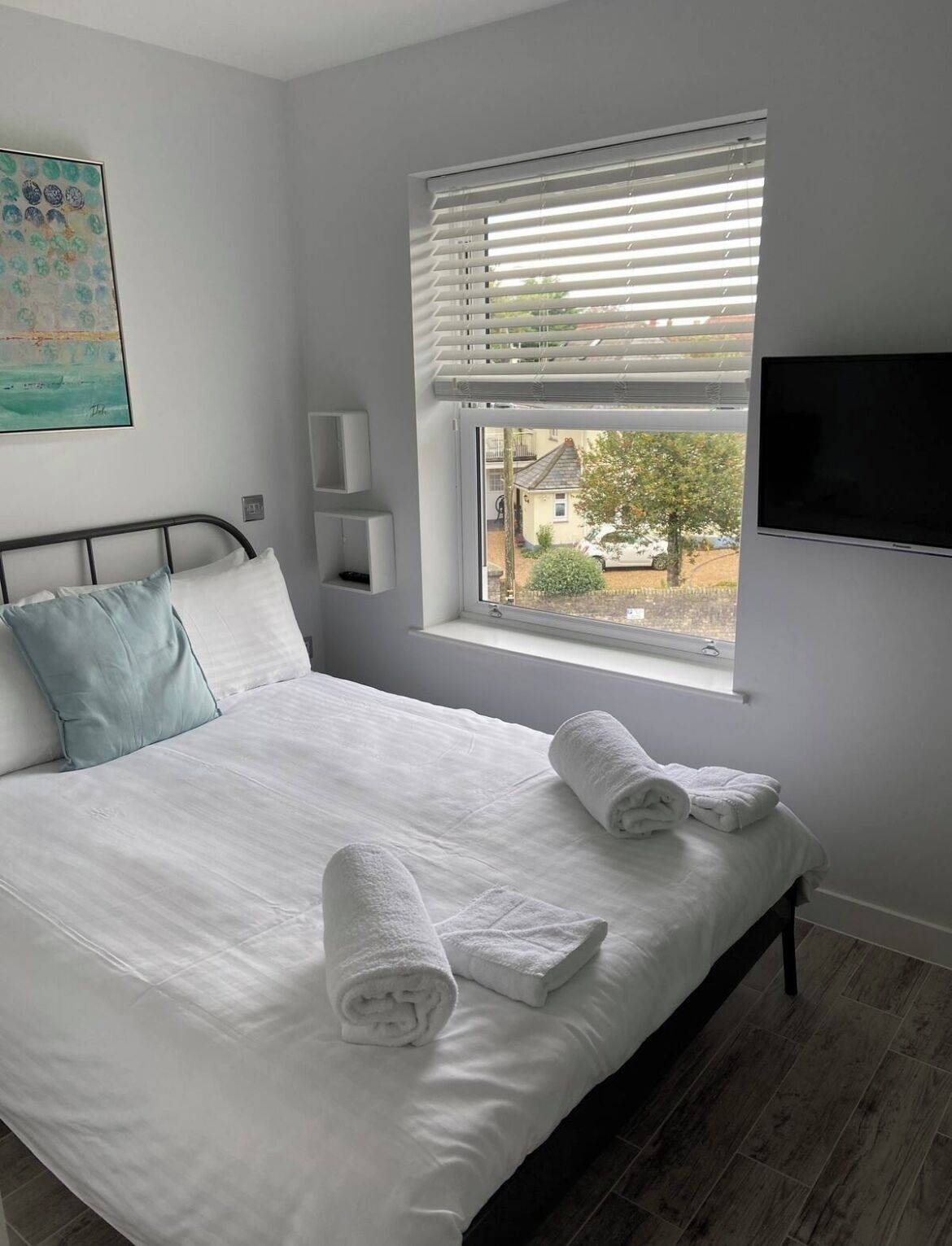 Une chambre à coucher avec un lit, une fenêtre avec des stores, un téléviseur à écran plat et un tableau accroché au mur.