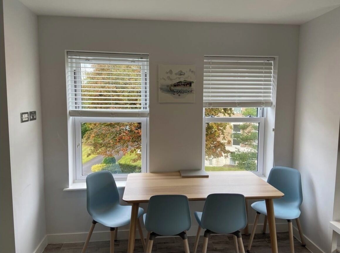 A dining area with a table and four chairs, two windows with blinds, and a framed picture on the wall.