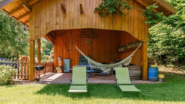 Een houten paviljoen met een hangmat, twee groene lounge fauteuils en een barbecue.