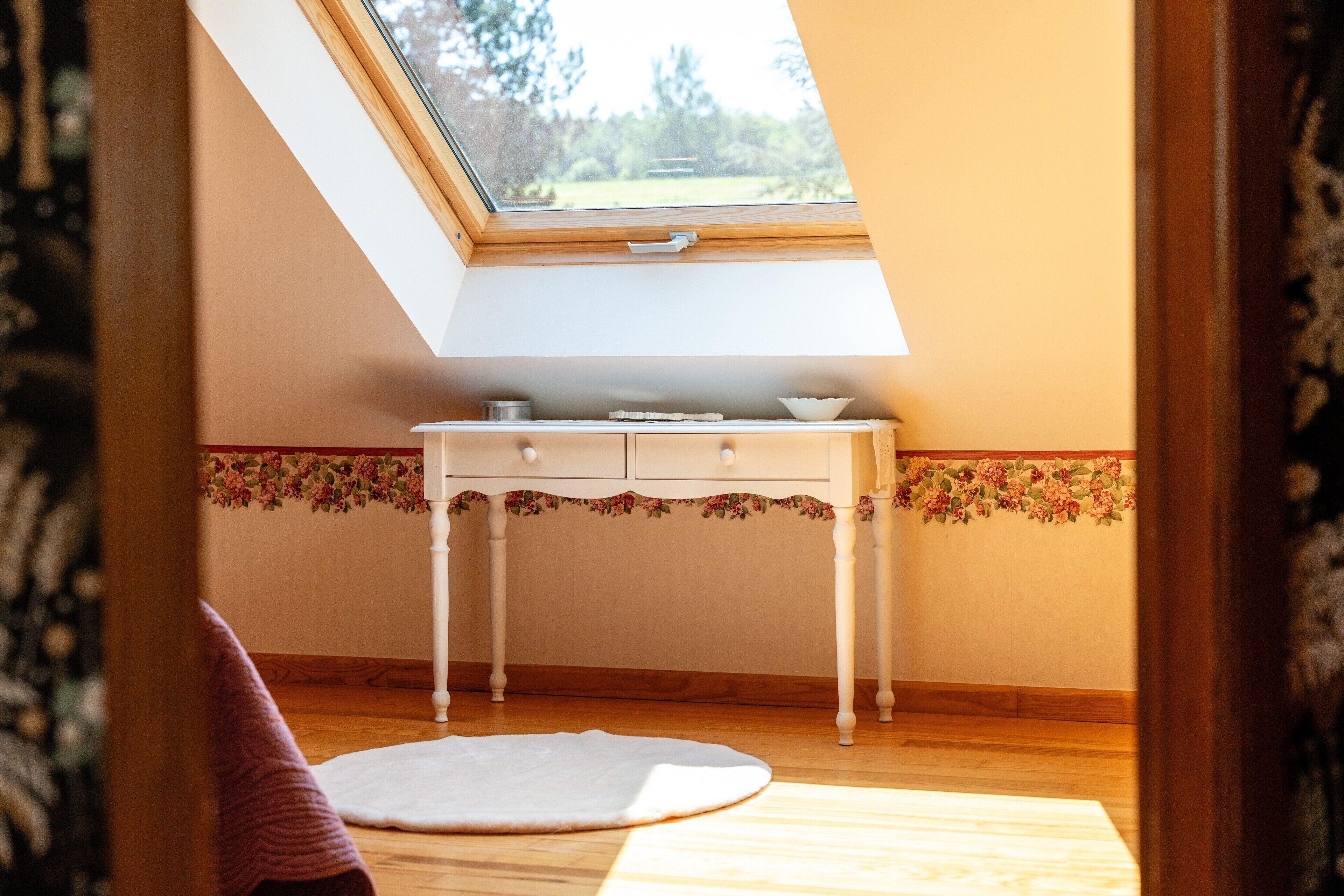 Een kamer met dakraam, een wit bureau met lades, een bloemenrand op de muur en een witte vloerkleed.