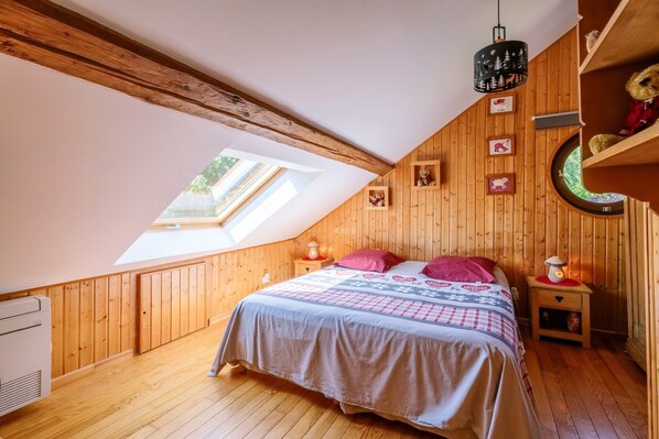 Une chambre avec un plafond en pente, des murs en bois, un lit recouvert d’une couvre-lit à motifs, une petite table de chevet avec une lampe et une fenêtre ronde.