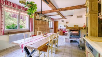 Una cocina rústica con una mesa de madera, sillas, lámparas colgantes, una chimenea y vistas al exterior con vegetación.