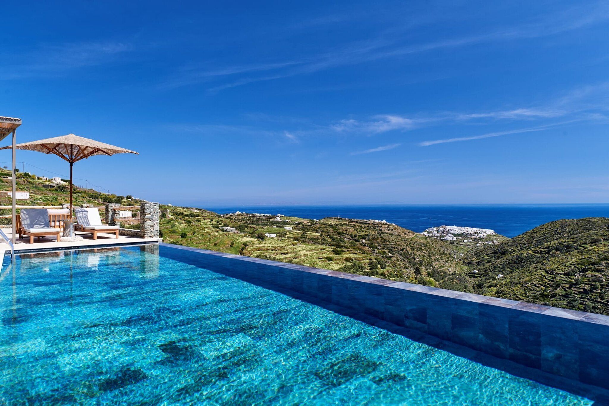 Una piscina azul cristalina con sillas de descanso y sombrillas, con vistas a un paisaje montañoso y al mar.