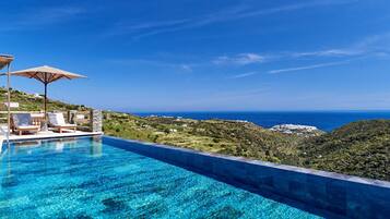 Una piscina azul cristalina con sillas de descanso y sombrillas, con vistas a un paisaje montañoso y al mar.