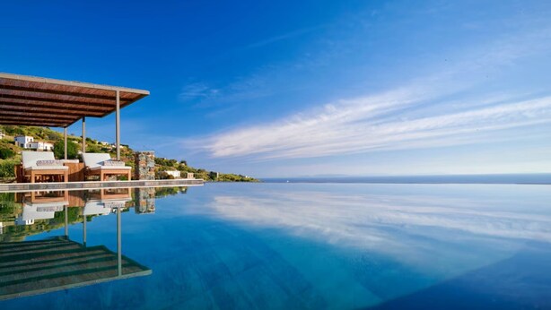 Vista chiara di una piscina a sfioro con un pontile in legno e poltrone prendisole, che riflette il paesaggio circostante e un orizzonte lontano.