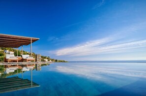 A clear view of an infinity pool with a wooden deck and lounge chairs, reflecting the surrounding landscape and a distant horizon.