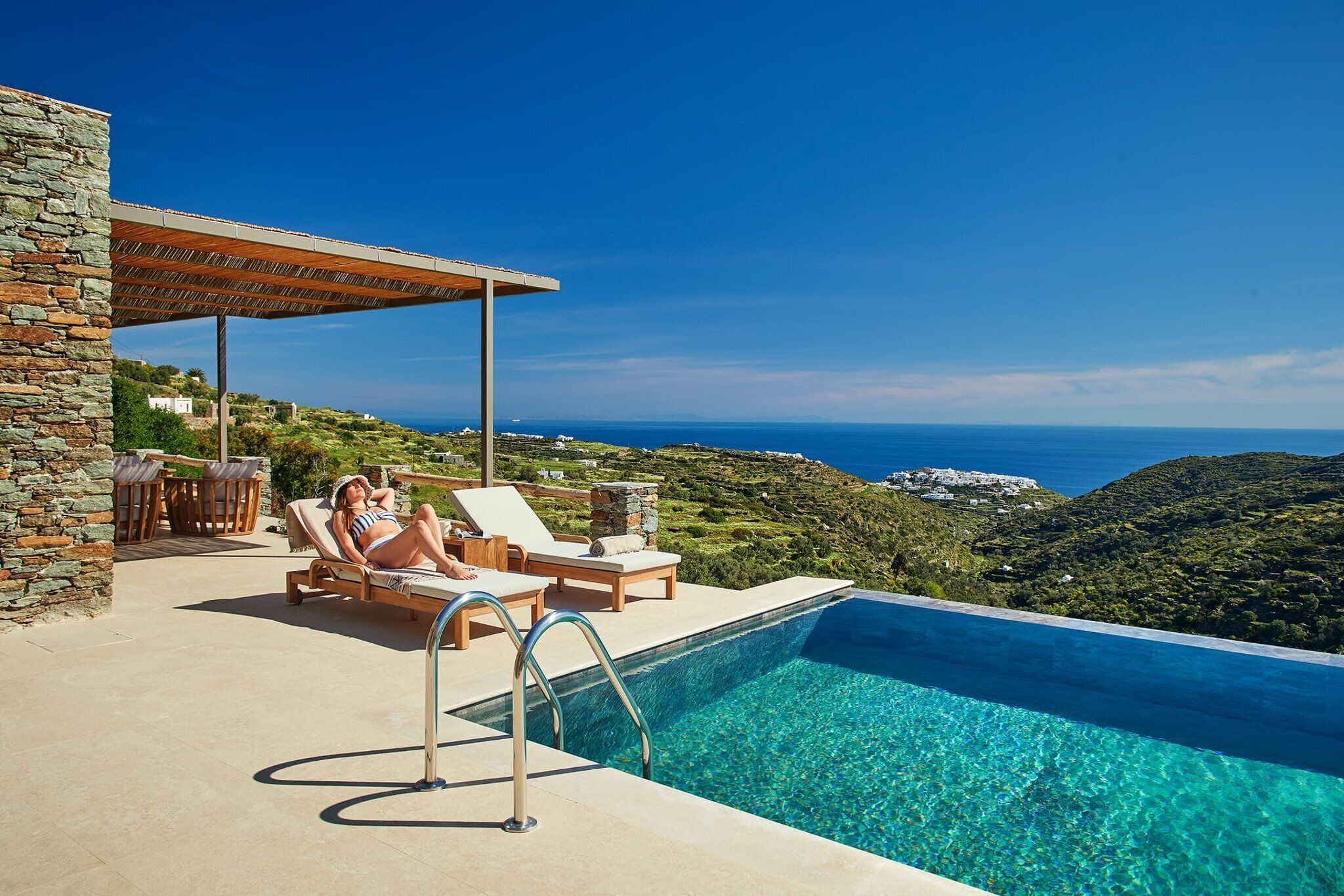 Zona de piscina con vista despejada al mar, pared de piedra y área de descanso cubierta.