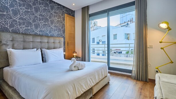 Ein modernes Hotelzimmer mit einem großen Bett, Blick auf die Stadt durch eine Glastür und einer auffälligen, gelben Lampe.