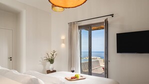 Hotelkamer met een bed, een flatscreen-tv, een glazen schuifdeur naar een balkon met uitzicht op zee, en een tafeltje met een glas wijn en fruit.