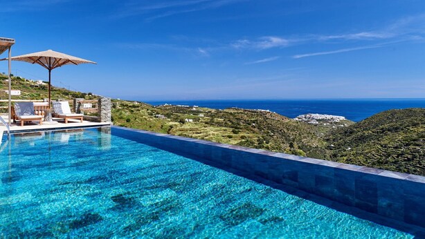 Una piscina azul cristalina con sillas de descanso y sombrillas, con vistas a un paisaje montañoso y al mar.