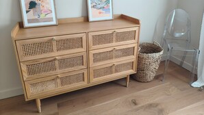 Une commode en bois avec des portes en osier, deux œuvres d’art encadrées posées sur le dessus, et une chaise en plastique transparent placée à côté.