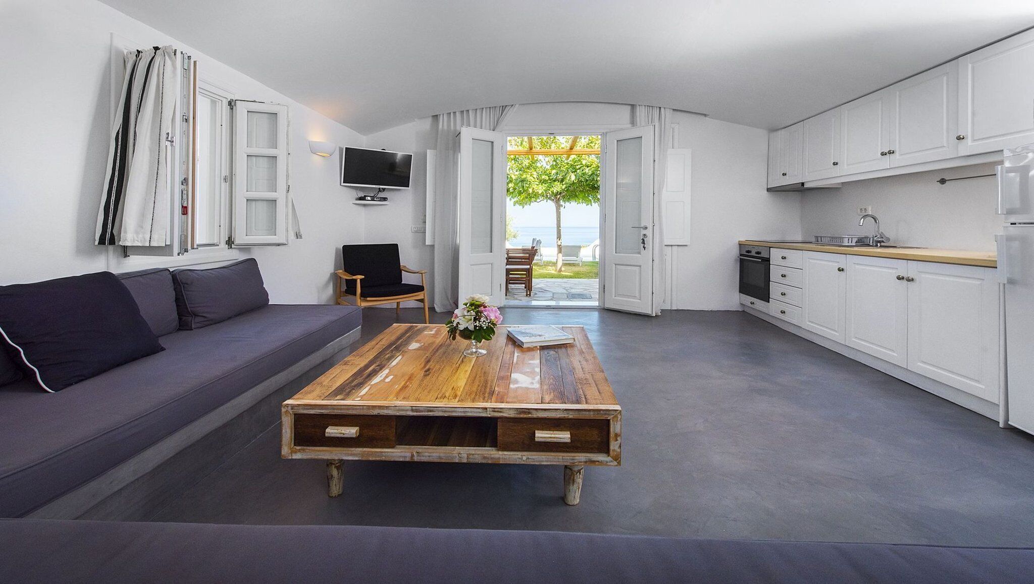 Ein modernes Wohnzimmer mit einer Couch, einem Holztisch und einem Fernseher. Ein Küchenbereich mit Schränken und ein Essbereich sind durch eine offene Tür zu sehen.