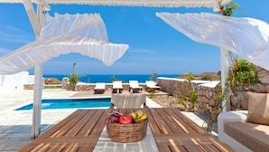 Un espace extérieur aménagé, abrité et doté d’une piscine et de chaises longues, offrant une vue dégagée sur un ciel bleu azur et l’océan.