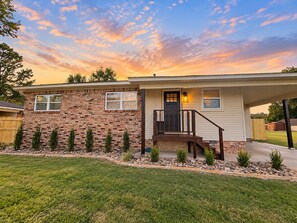 A two-story house with a brick facade, a front porch with a staircase, and a well-maintained lawn. - The Loves’ Shack With Fire Pit, EV Charger, near I40, AR Nuclear 1 & Data Center (Clarksville)