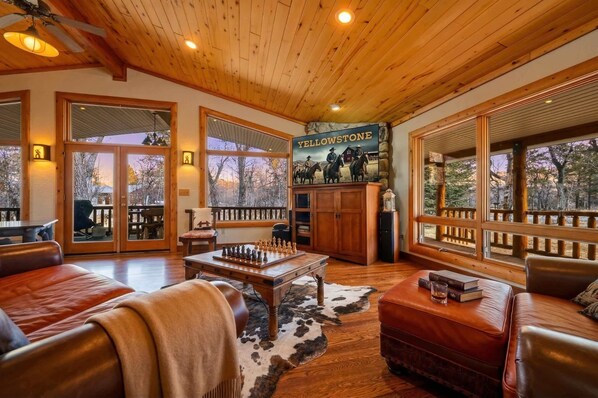 A spacious living room with wooden flooring, leather furniture, a mounted chessboard, and large windows offering a view of the outdoors. - Bunkhouse — Ranch Estate, Private Pond, Story WY (Banner)