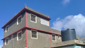 Un bâtiment à deux étages, doté d’un toit plat, d’une citerne d’eau sur le côté droit et d’une petite structure adjacente avec une porte et une fenêtre.