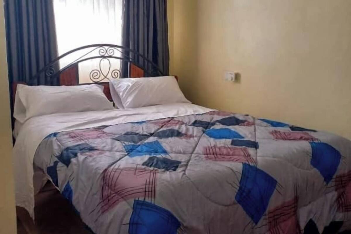 Ein Einzelbett mit einem gemusterten Bettbezug, ein Fenster mit blauen Vorhängen und eine schlichte Wand.