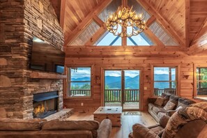 A log cabin interior with a stone fireplace, wooden furniture, and large windows offering mountain views. - Waynesville Mtn Cabin-scenic Views Relaxing Hottub (Waynesville)